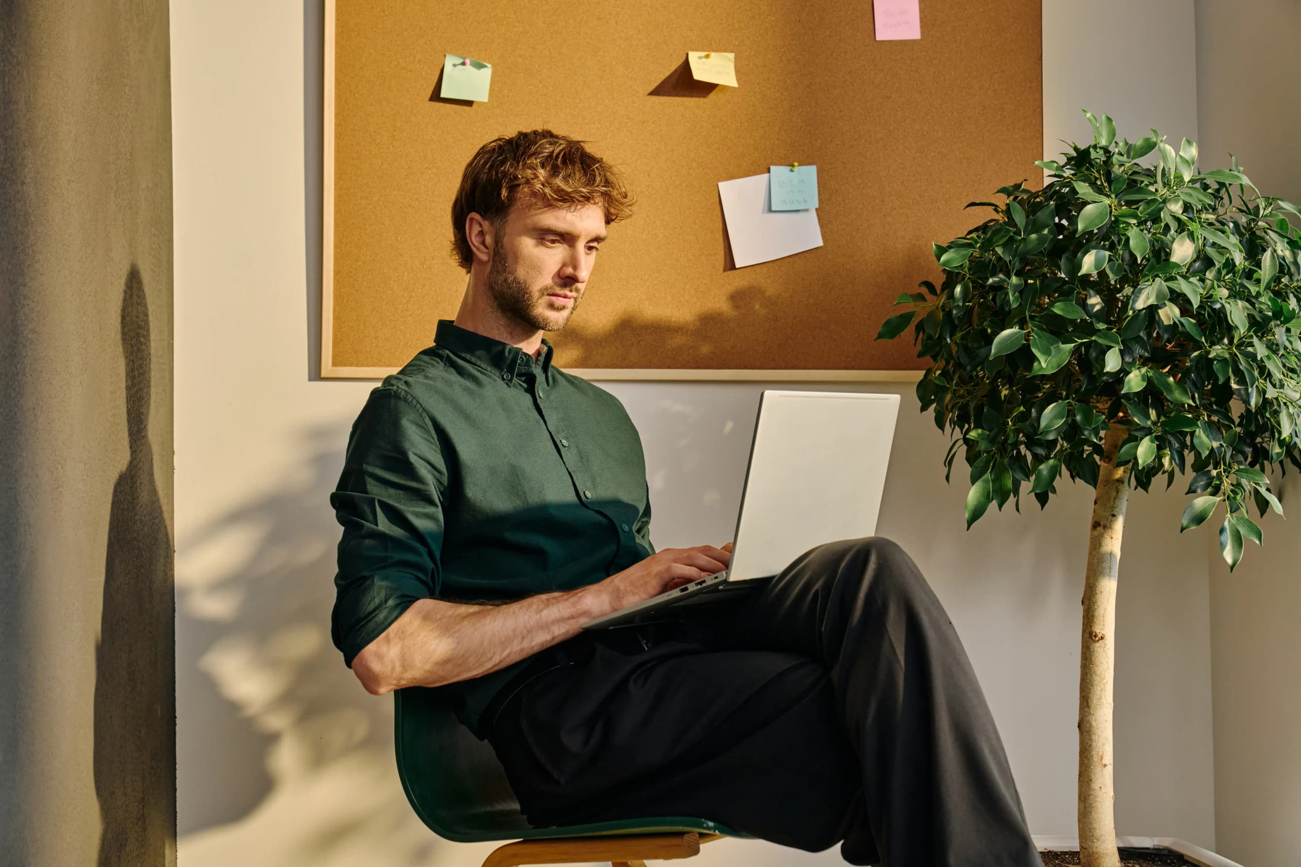 Чоловік сидить на стільці з ноутбуком на колінах біля дошки з наліпками та поруч із кімнатною рослиною Чоловік сидить на стільці з ноутбуком на колінах біля дошки з наліпками та поруч із кімнатною рослиною