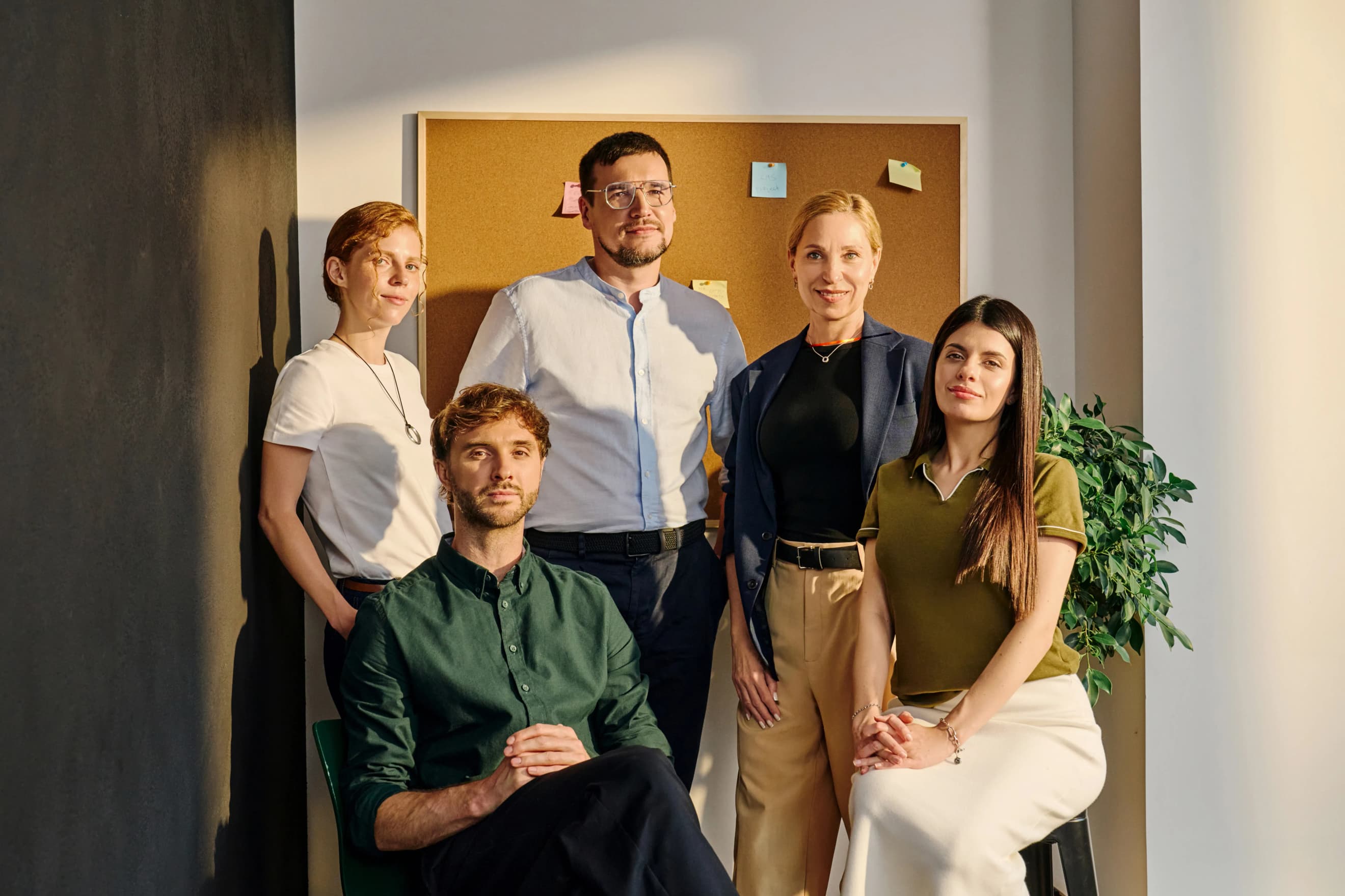 Група людей позує для фото в офісі перед корковою дошкою з різнокольоровими наліпками Група людей позує для фото в офісі перед корковою дошкою з різнокольоровими наліпками