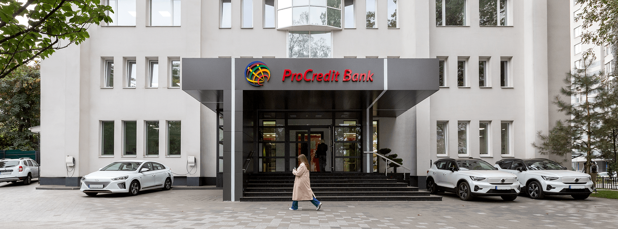 Вхід до ПроКредит Банку із темно-сірим навісом, сходами та скляними дверима, поруч припарковані електроавтомобілі Вхід до ПроКредит Банку із темно-сірим навісом, сходами та скляними дверима, поруч припарковані електроавтомобілі