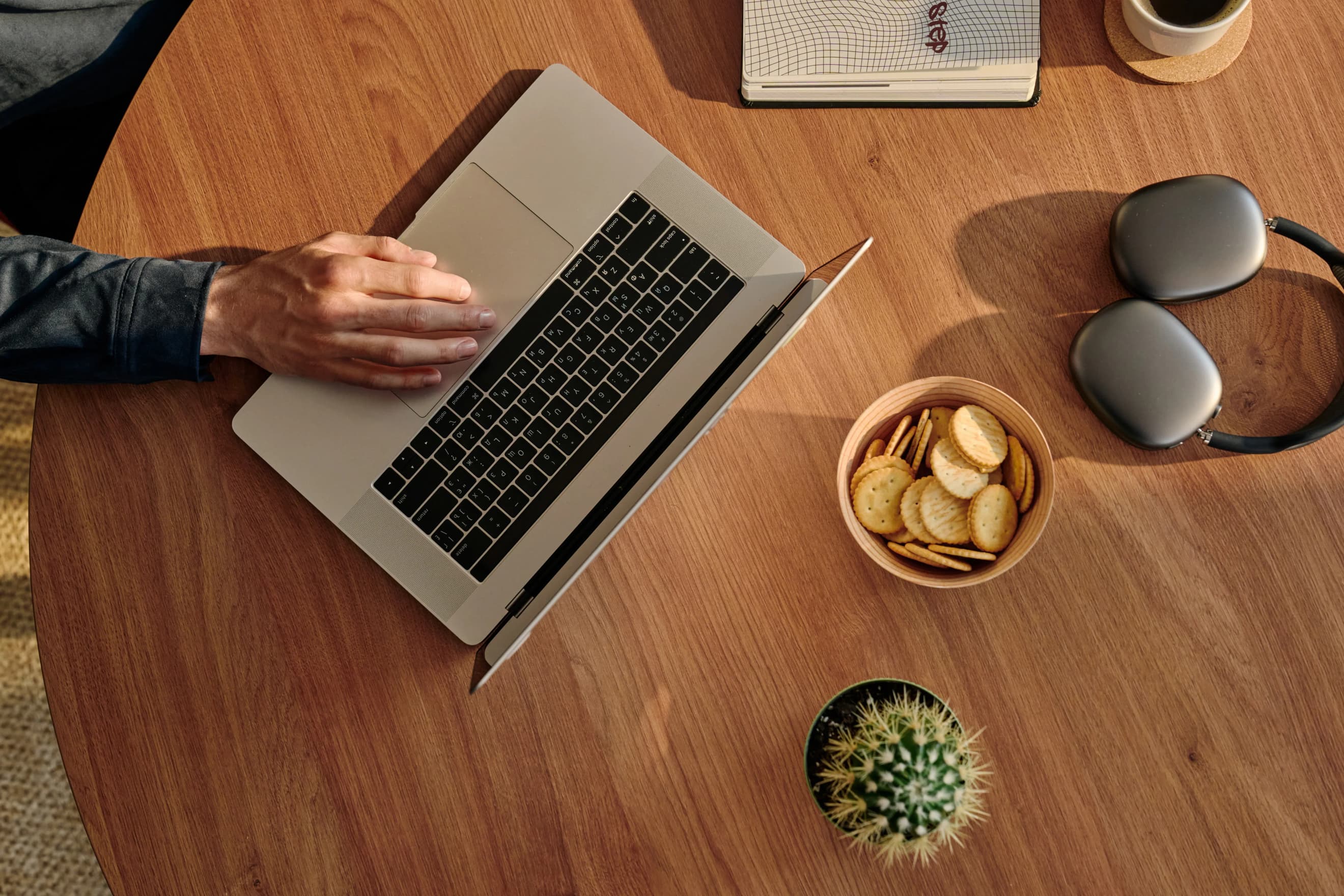 Рука працює за ноутбуком на дерев’яному столі поруч із навушниками, блокнотом, кактусом і тарілкою з крекерами. Рука працює за ноутбуком на дерев’яному столі поруч із навушниками, блокнотом, кактусом і тарілкою з крекерами.
